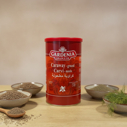A red canister of Gardenia Caraway ground carvi-moutarde spice is displayed on a light wooden surface, surrounded by small bowls containing whole seeds, a wooden spoon with seeds, and fresh dill, against a neutral beige background.